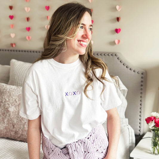 young woman wearing a white tee with two tones purple xoxo embroidered small on the center chest