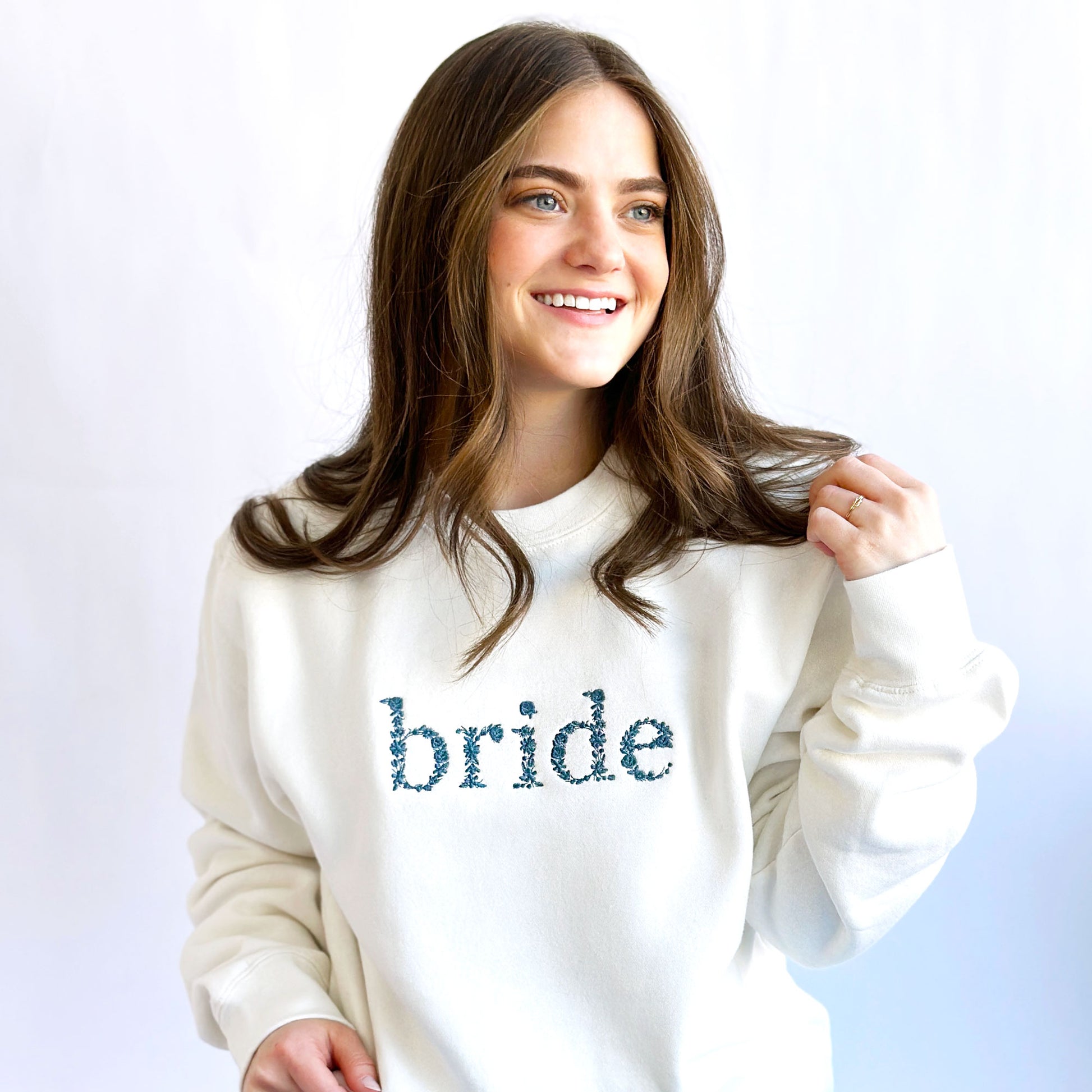 woman wearing a white crewnecksweatshirt with floral lowercase bride embroidered in french blue thread across the chest
