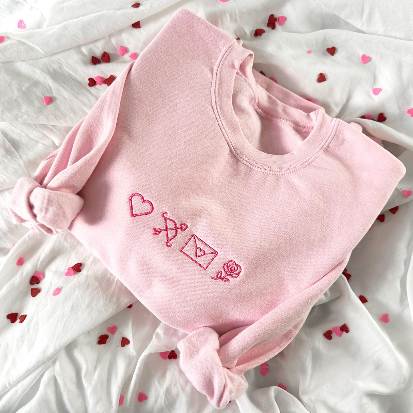 light pink crewneck sweatshirt with a heart, cupids bow and arroe, heart envelope, and a rose embroidered in pink across the chest