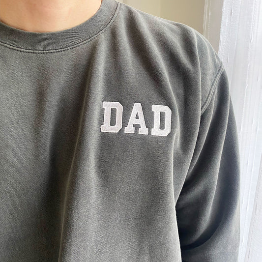 Close up of a man wearing a black crewneck sweatshirt with dad embroidered in white on the left chest