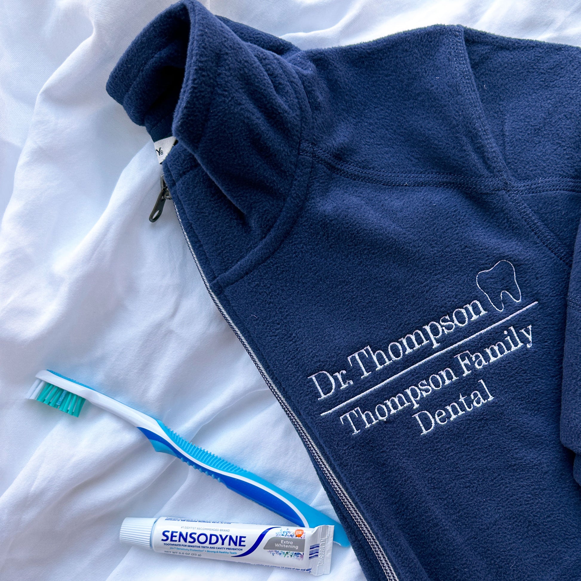 close up of a full zip navy fleece jacket with Dr, thompson Thompson family dental and mini outline tooth embroidered on the left chest on white thread