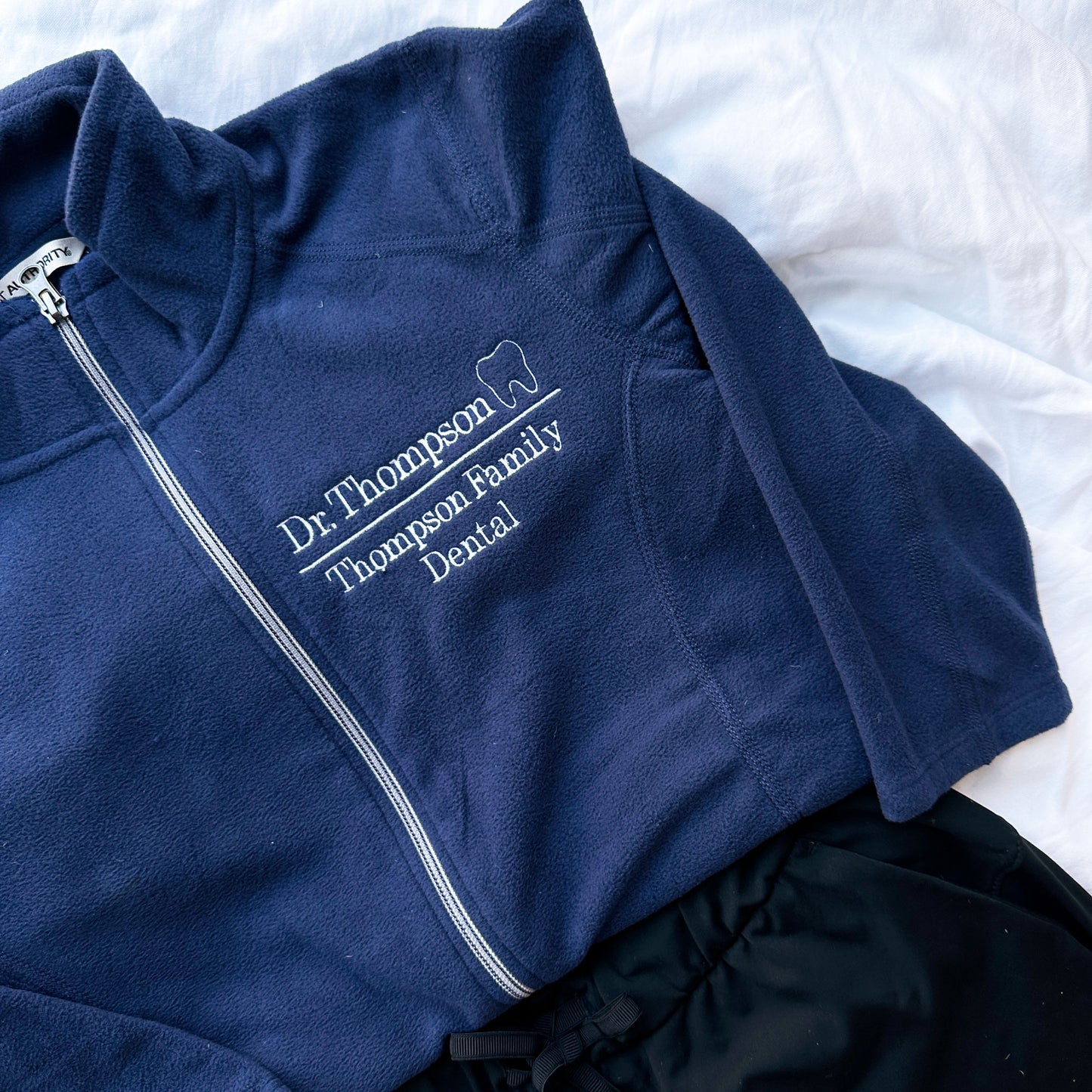close up of a full zip navy fleece jacket with Dr, thompson Thompson family dental and mini outline tooth embroidered on the left chest on white thread