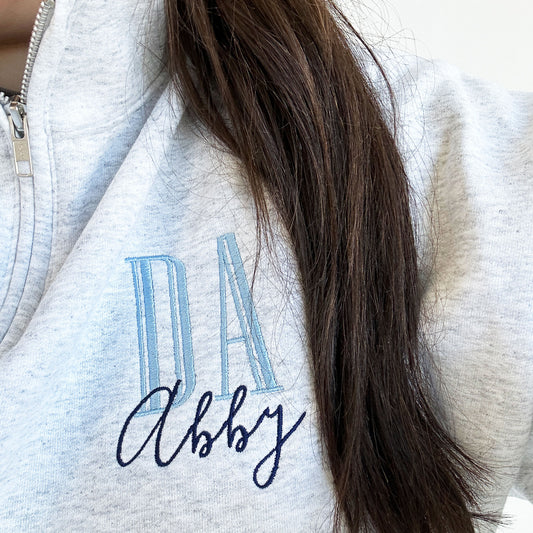 close up of an embroidered ash quarterzip  sweatshirt with DA in baby blue thread and large outline letters and name Abby below DA in navy thread and a loose script font