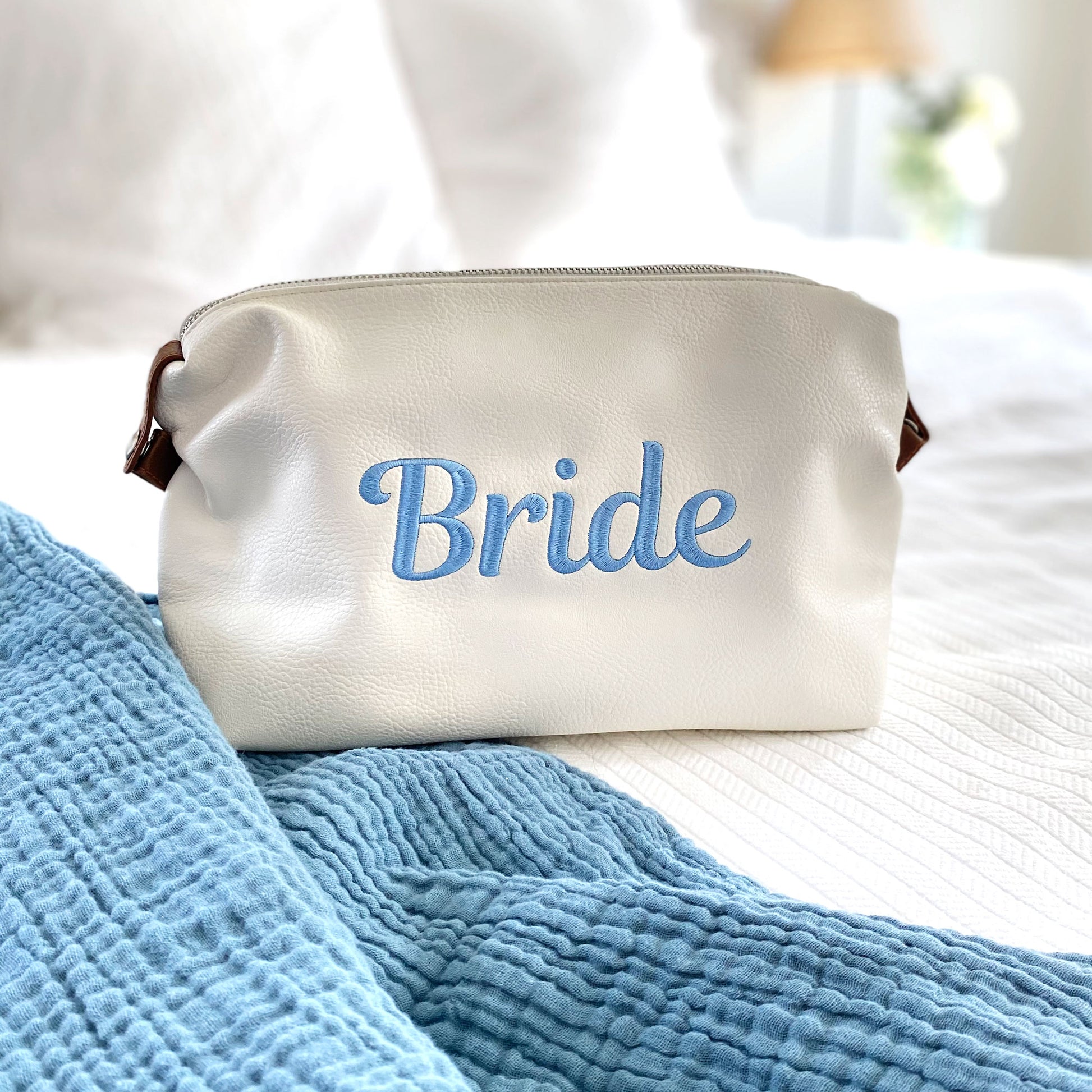 White addison dopp kit with script bride embroidered in baby blue thread on the center of the bag styled on a bed