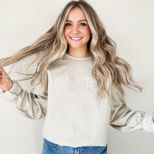 bride to be wearing a sand colored sweatshirt with wifey embroidered in a varstiy font across the chest
