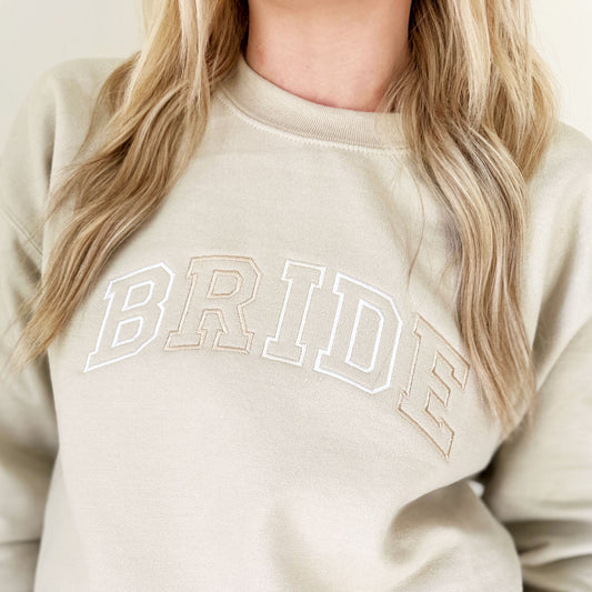 close up of a BRIDE design embroidered in an atheltic block open font across the chest of a sand sweatshirt in various neutral thread colors