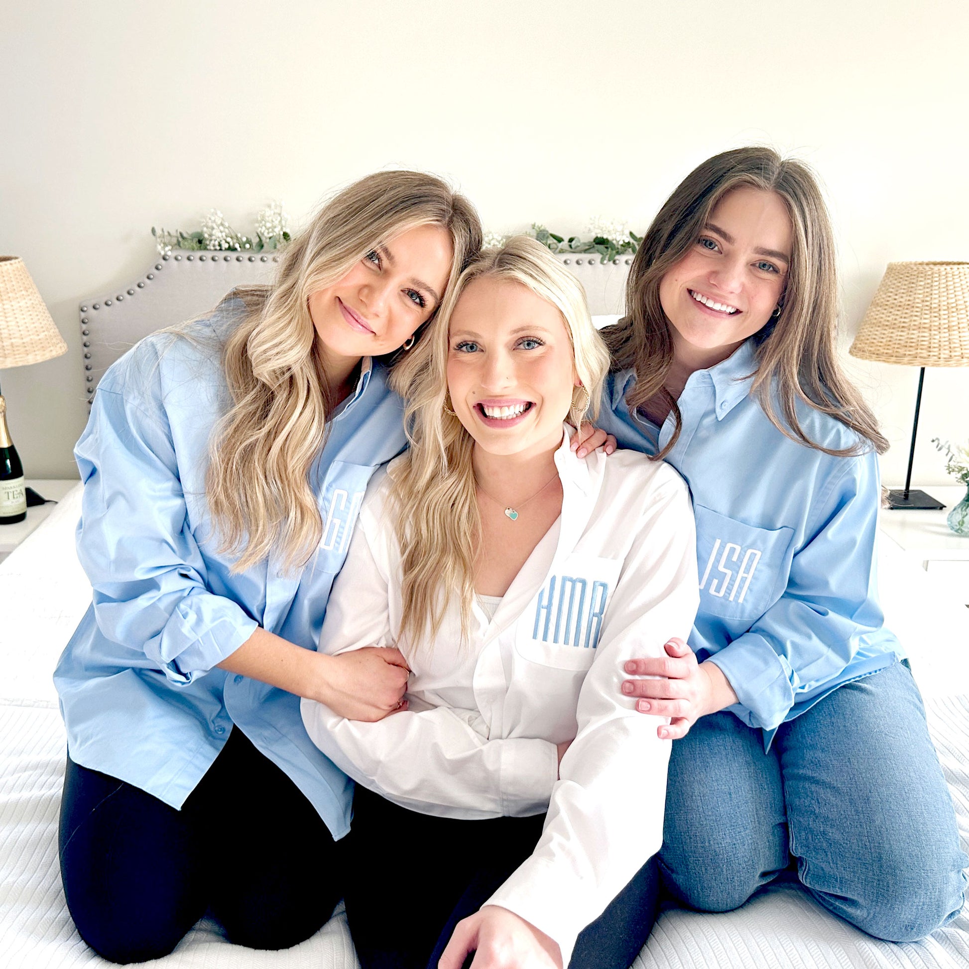 bride and 2 bridesmaids modeling white and light blue button down shirts with font 18 moogram embroidered on the left chest pocket in white and baby blue threads
