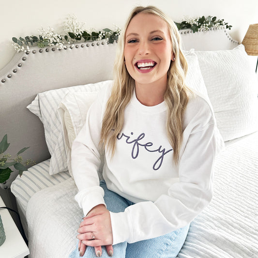 Young woman wearing a white crewneck sweatshirt with wifey embroidered across the chest in smoky orchid thread