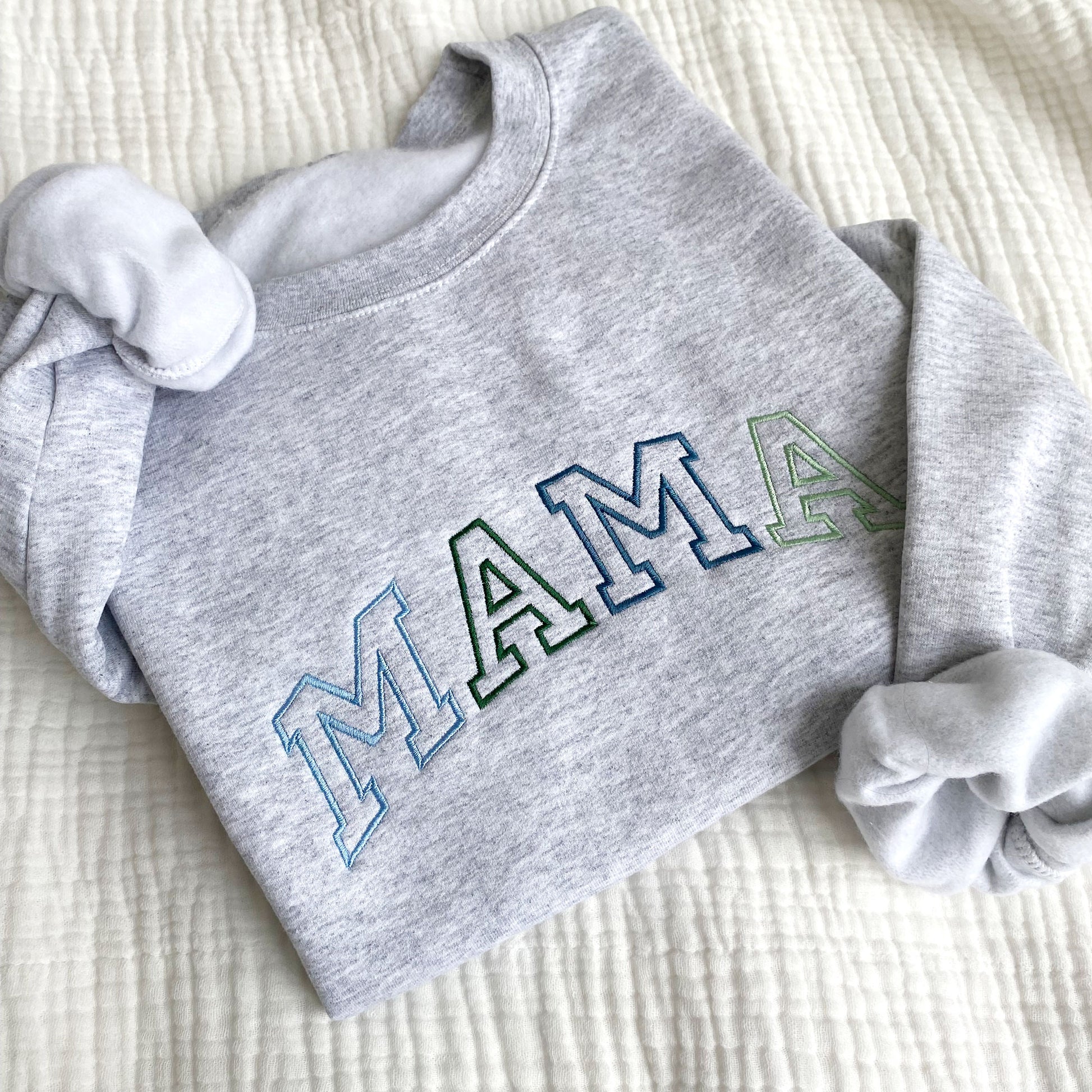 flat lay photo of an ash crewneck sweatshirt with large embroidered athletic block MAMA in alternating blues and greens