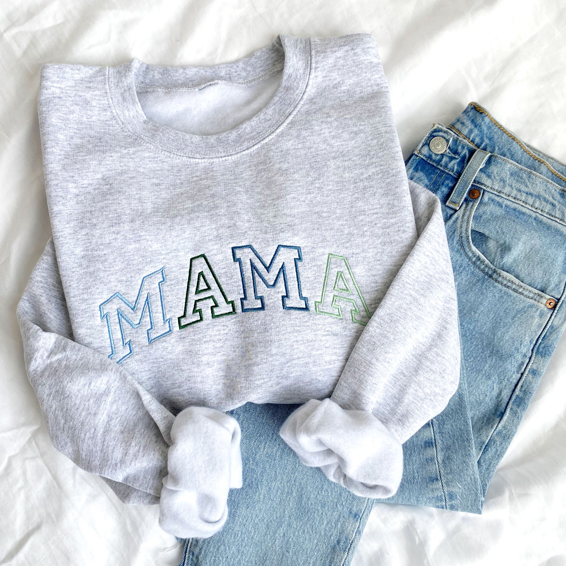 flat lay photo of an ash crewneck sweatshirt with large embroidered athletic block MAMA in alternating blues and greens styled with jeans