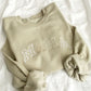 flat lay photo of a sand crewneck sweatshirt with embroidered MAMA in alternating neutral threads across the chest