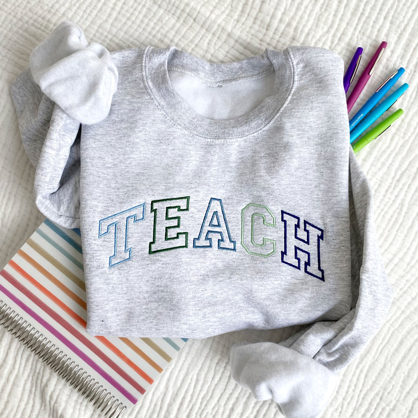flat lay photo of an ash crewneck sweatshirt with athletic block outline embroidered TEACH in blues and greens