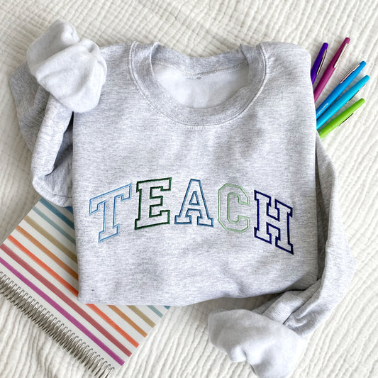 flat lay photo of an ash crewneck sweatshirt with athletic block outline embroidered TEACH in blues and greens