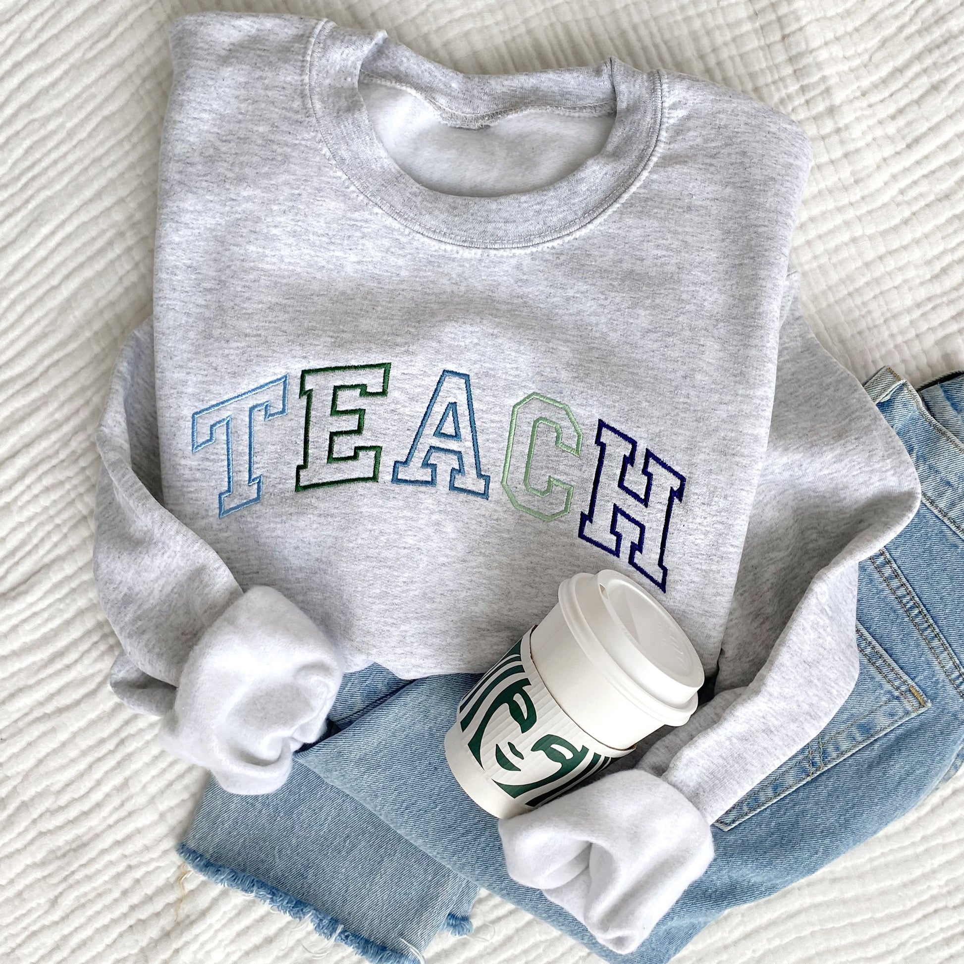 flat lay photo of an ash crewneck sweatshirt with athletic block outline embroidered TEACH in blues and greens