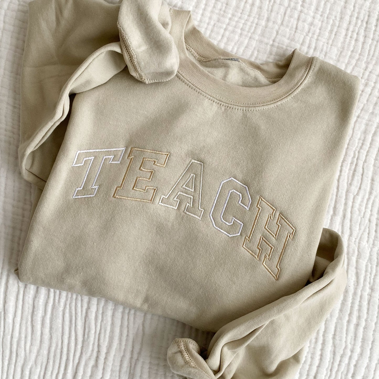 flat lay of a sand crewneck sweatshirt with embroidered neutral color block teach design across the chest
