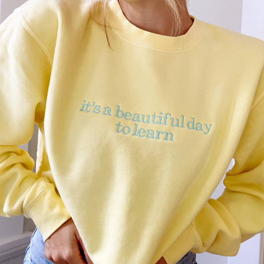 GIRL WEARING YELLOW SWEATSHIRT WITH CUSTOM EMBROIDERY READING 'IT'S A BEAUTIFUL DAY TO LEARN' IN A SKY BLUE THREAD