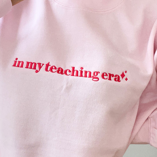 close up of a a light pink crewneck sweatshirt with embroidered in m teaching era design across the chest in pink thread