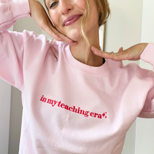 young woman wearing a light pink crewneck sweatshirt with embroidered in m teaching era design across the chest in pink thread