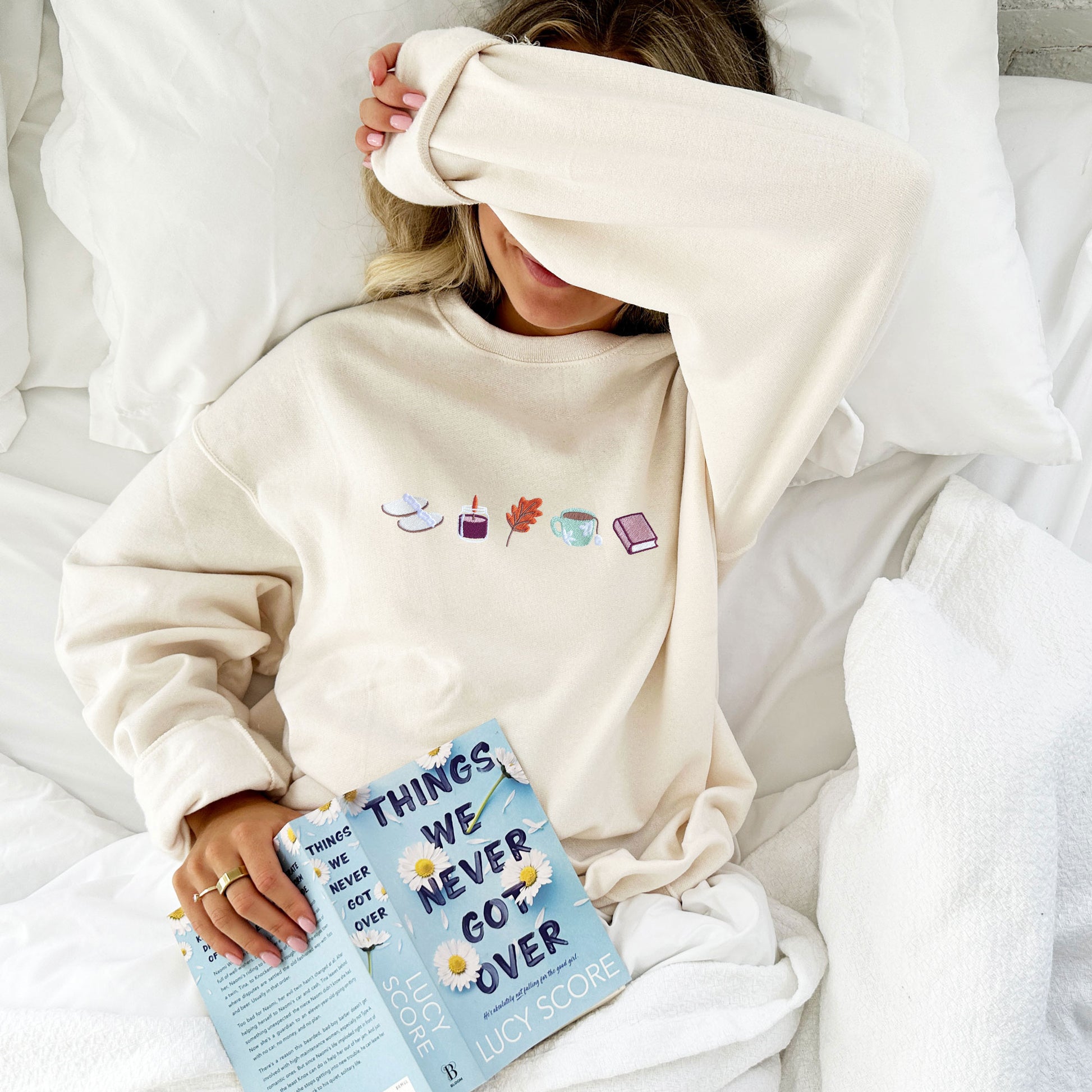 woman laying on a bed modeling an oversized sweatshirt with embroidered fall icons across the chest
