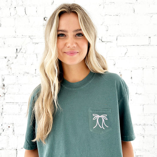 girl wearing a blue spruce pocket tee with a cute coquette ribbon bow embroidered design on the left chest pocket