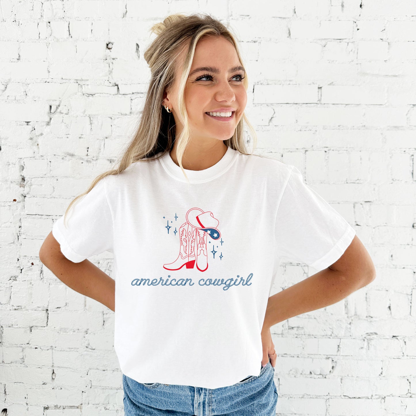 girl wearing an oversized white comfort colors t-shirt with a cute american cowgirl print in a rope font with cowgirl boots and a cowgirl hat
