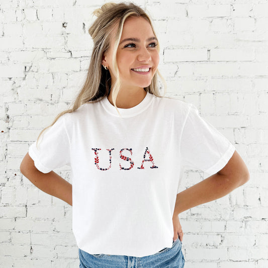 young woman modeling a white comfort colors t-shirt with embroidered floral USA design in red, white, and navy threads