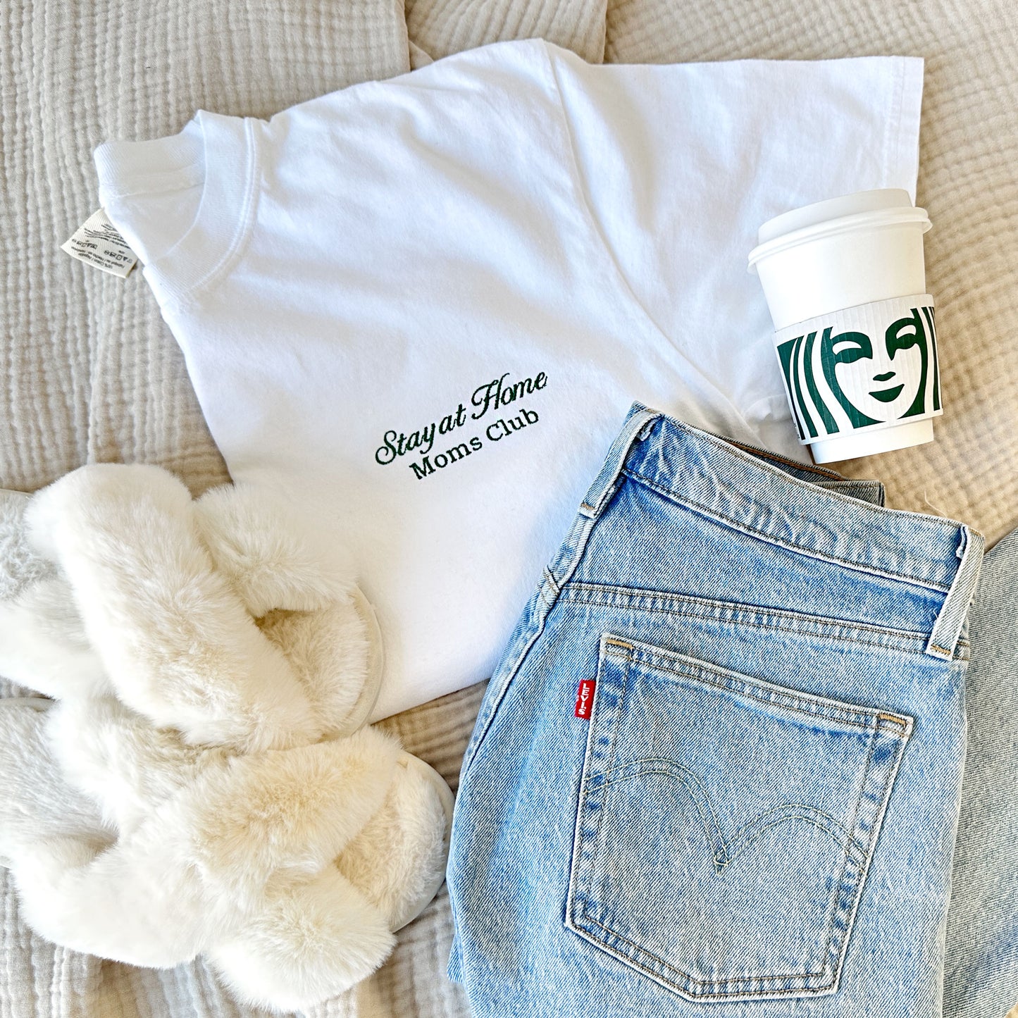 styled flat lay of a white comfort colors tshirt with embroidered stay at home moms club design small on the left chest in hunter green thread with jeans, slippers, and a starbucks cup