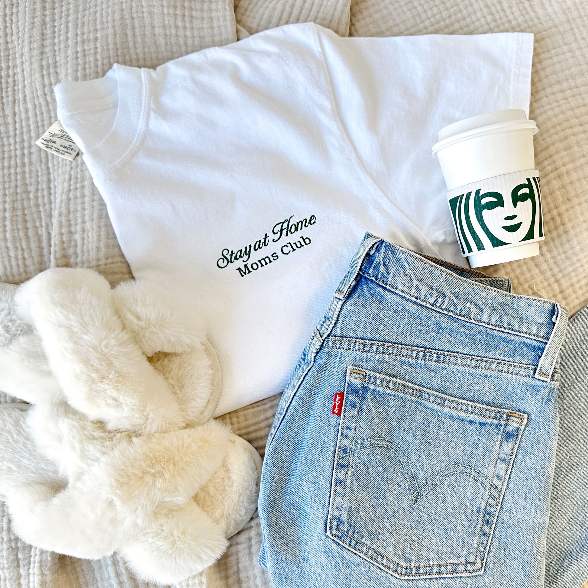 styled flat lay of a white comfort colors tshirt with embroidered stay at home moms club design small on the left chest in hunter green thread with jeans, slippers, and a starbucks cup