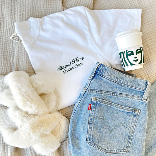 styled flat lay of a white comfort colors tshirt with embroidered stay at home moms club design small on the left chest in hunter green thread with jeans, slippers, and a starbucks cup