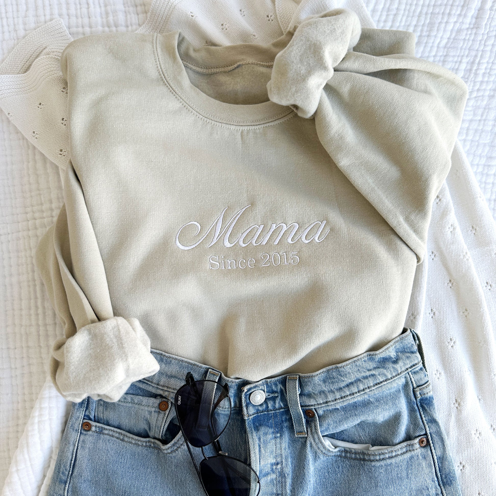 styled flat lay of a sand crewneck sweatshirt with embroidered Mama since customizable date in white thread across the chest with jeans and sunglasses