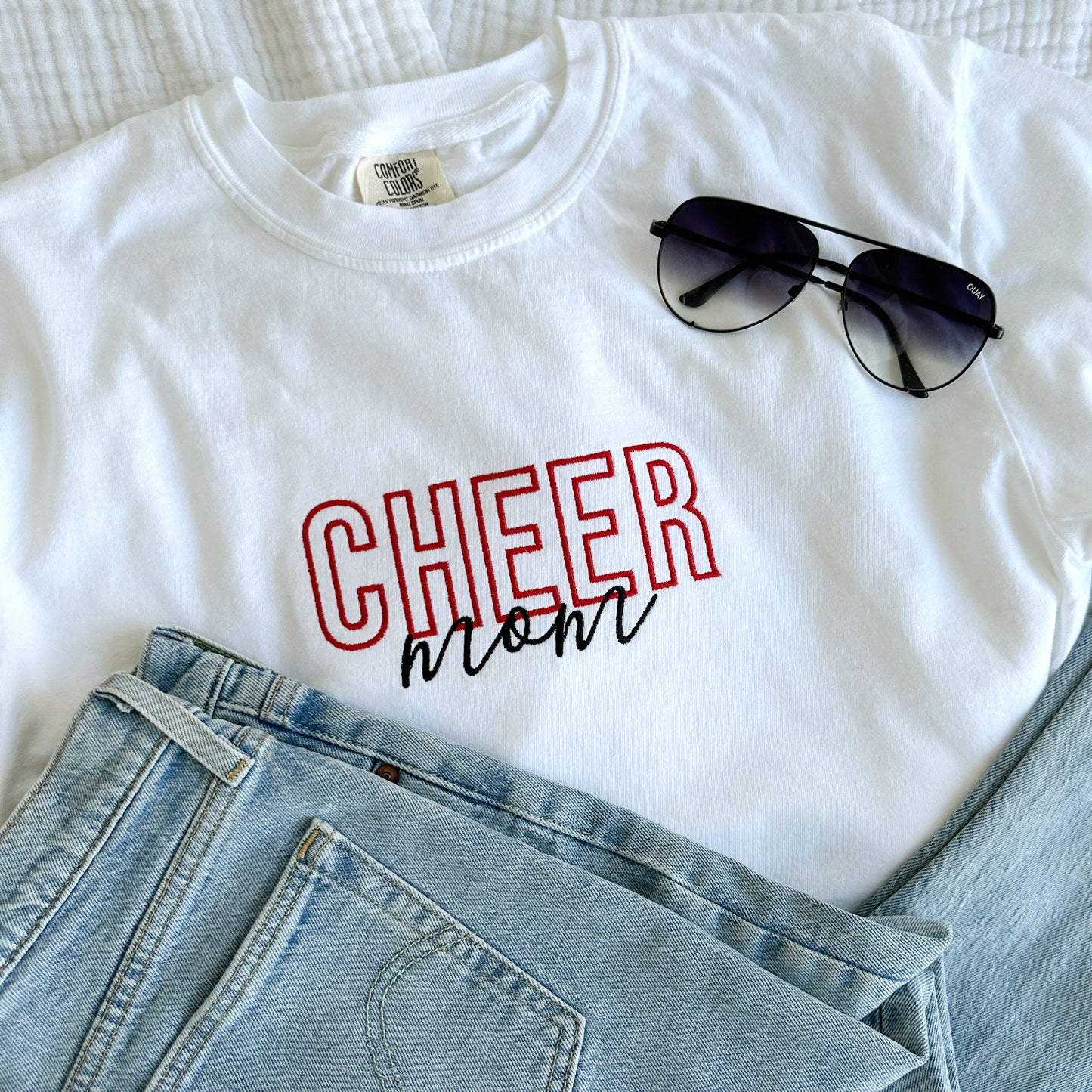 styled flat lay of a white comfort colors t-shirt with embroidered Cheer Mom design in black and red threads across the chest with jeans and sunglasses