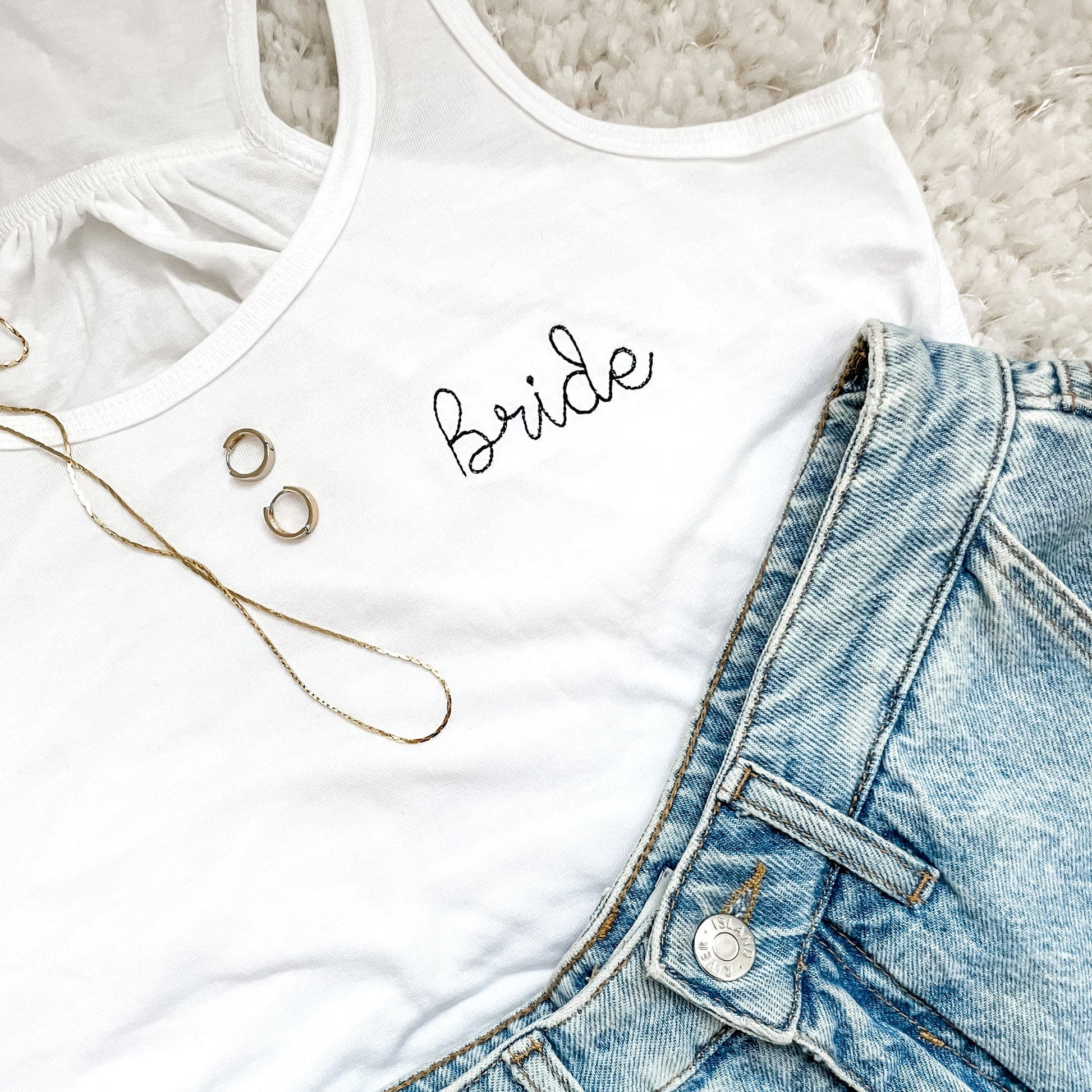 flat lay of white flowy bella and canvas tank top with bride embroidered in black thread paired with jewelry and jean shorts 
