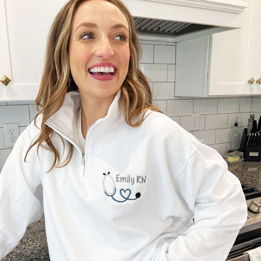up close of a woman wearing a white quarter zip pullover sweatshirt with a custom name and mini heart stethoscope embroidered design on the left chest.