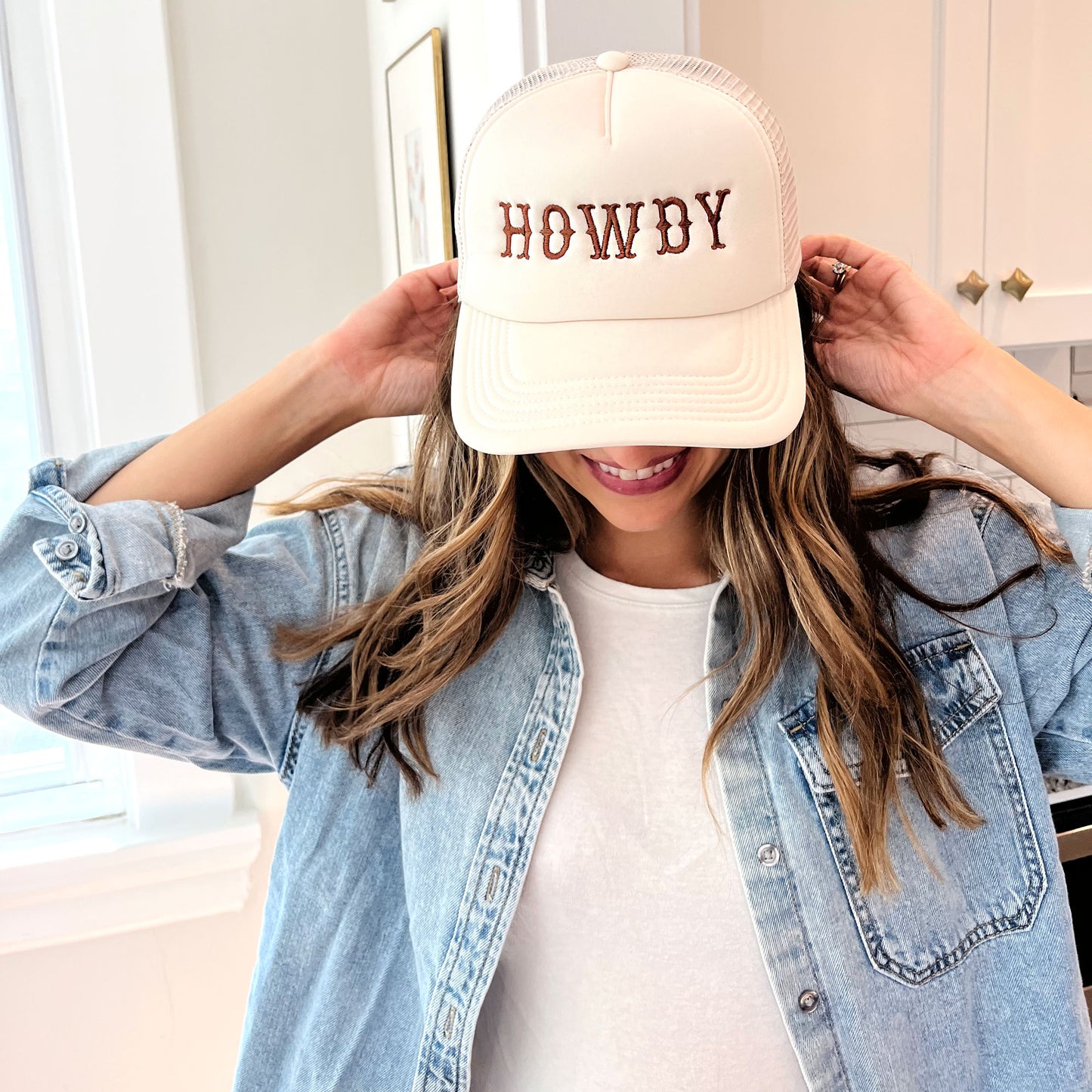 woman wearing a sand trucker hat with embroidered all caps howdy design in brown thread