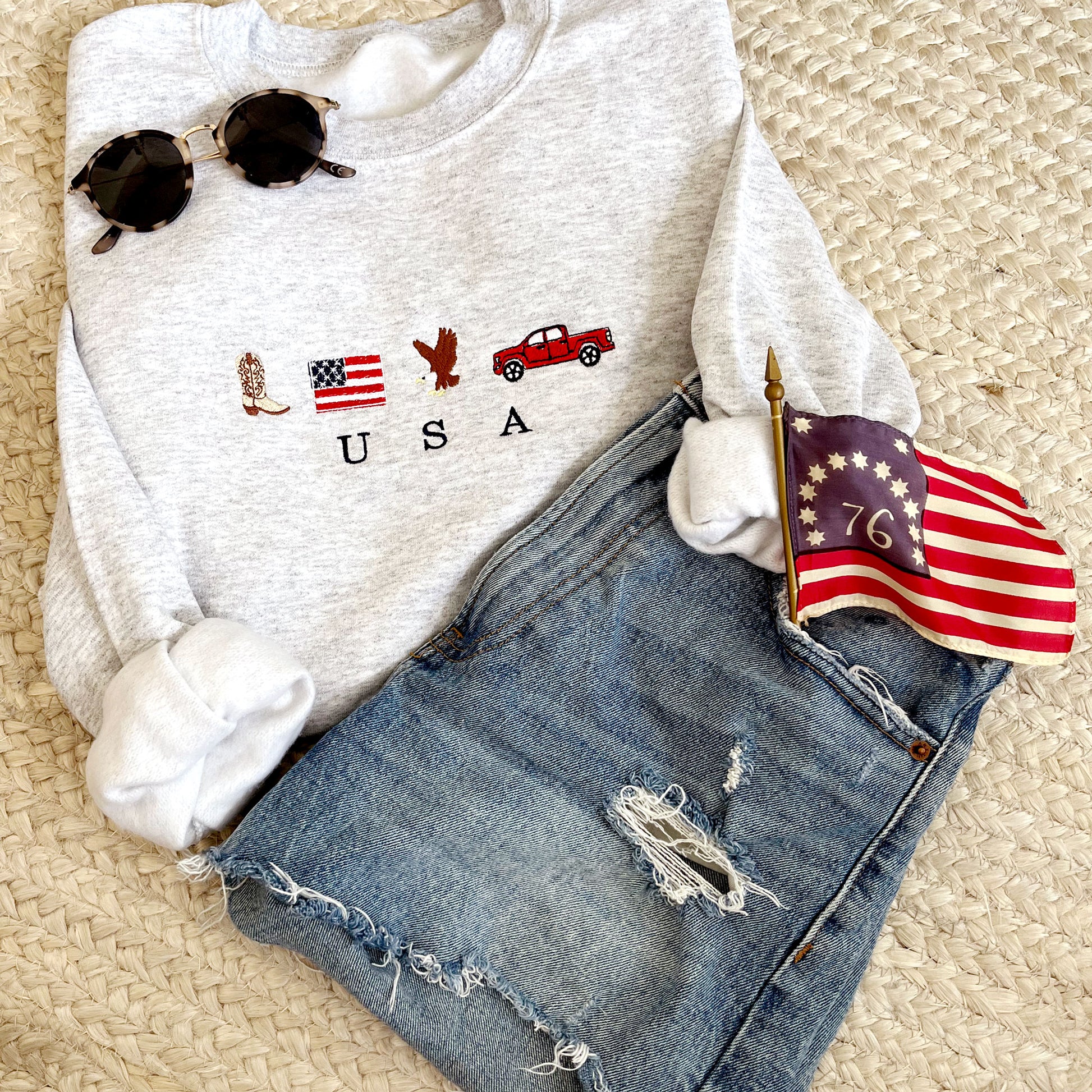 ASH GREY CREWNECK SWEATSHIRT WITH EMBROIDERED COWBOY BOOT, AMERICAN FLAG, EAGLE, TRUCK AND USA EMBROIDERED DESIGN ACROSS THE CENTER CHEST PAIRED WITH JEAN SHORTS