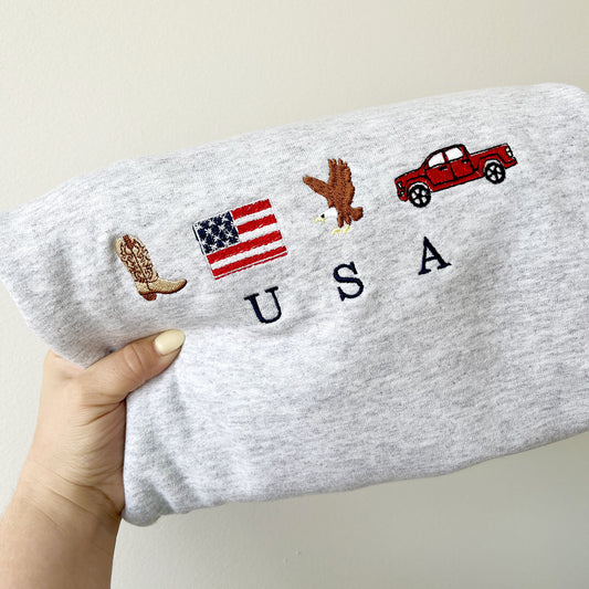 CLOSEUP OF AN ASH GREY CREWNECK SWEATSHIRT WITH EMBROIDERED COWBOY BOOT, AMERICAN FLAG, EAGLE, TRUCK AND USA EMBROIDERED DESIGN ACROSS THE CENTER CHEST