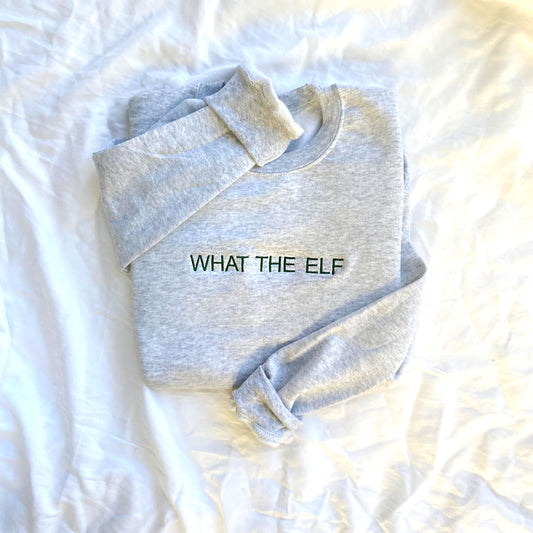 flat lay image of an ash gray sweatshirt with WHAT THE ELF embroidered across the chest in a dark green thread
