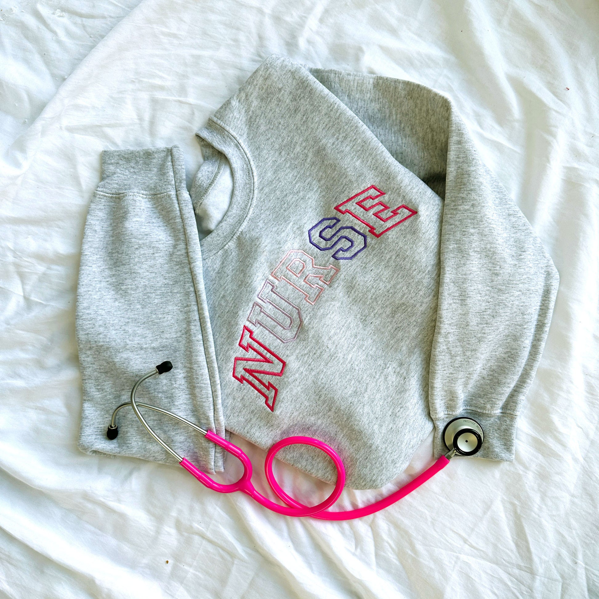 flat lay photo of an ash crewneck sweatshirt with embroidered athletic block nurse curved across the chest in alternating shades of pink and purple threads