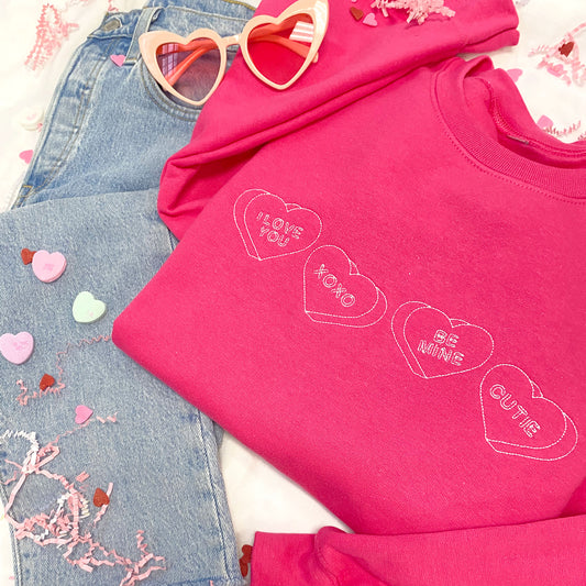 flat lay image of jeans, heart shaped glasses, a bright pink crewneck sweatshirt with stitched candy hearts across the chest  with "I love you," "xoxo," "be mine," and "cutie" designs