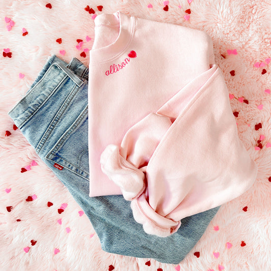 flat lay of blue jeans and a pink crewneck sweatshirt with a personalized name and mini heart embroidery along the neckline