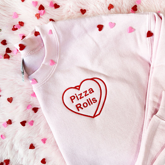 Flat lay image of a pink crewneck sweatshirt with a candy heart embroidered design that says pizza rolls in red thread