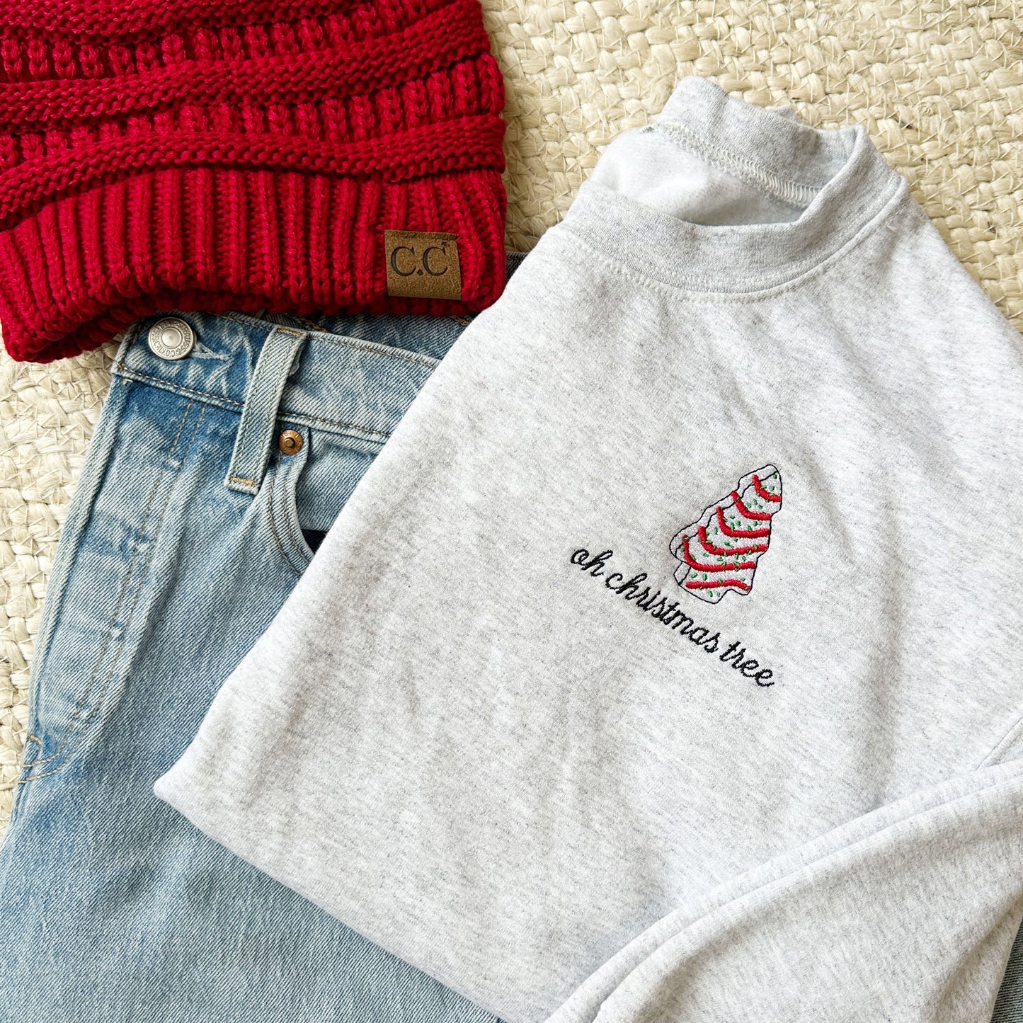Flat lay of an ash crewneck sweatshirt with a red beanie and blue jeans. On the left chest of the sweatshirt there is an embroidered little debbie christmas tree and the text oh Christmas tree below.