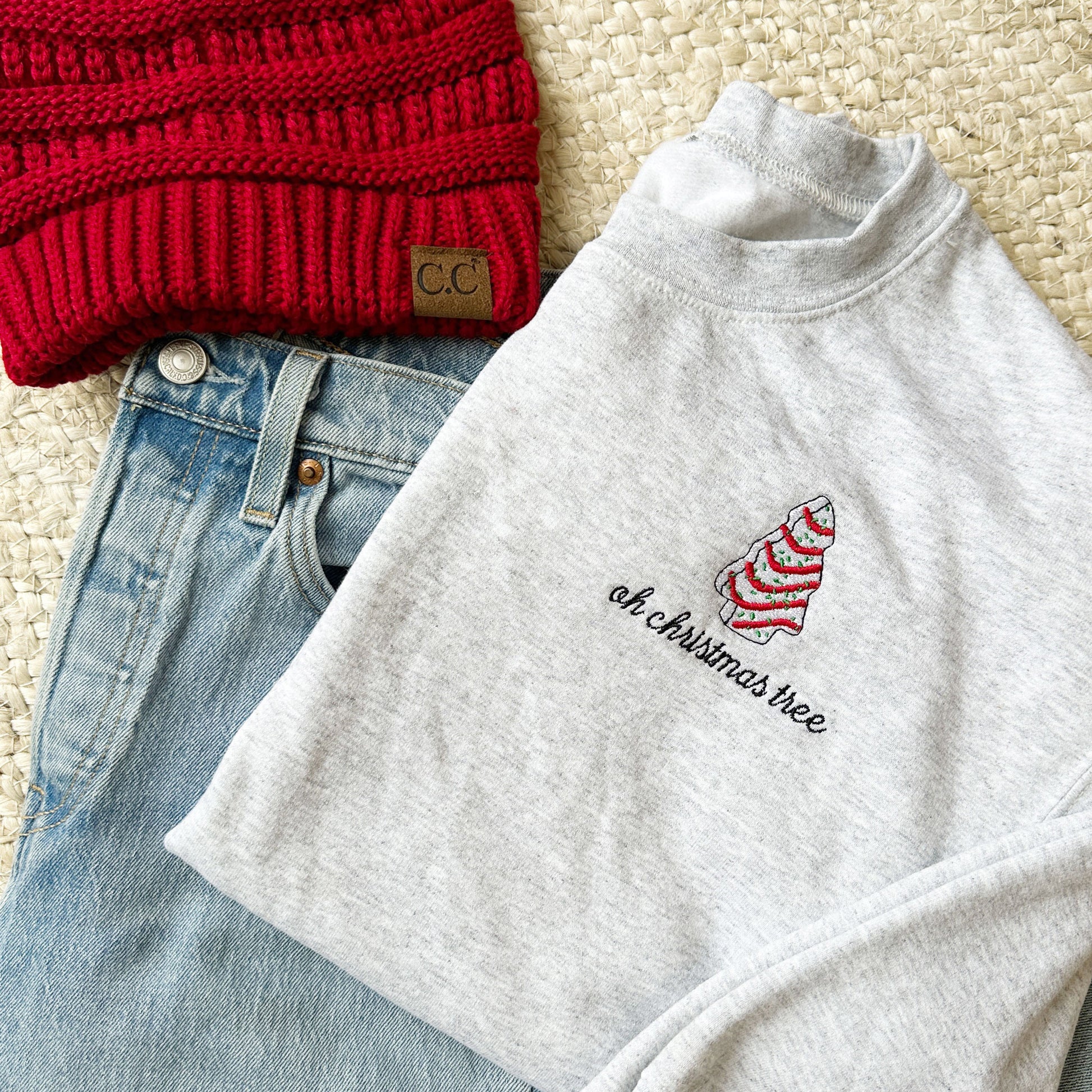 Flat lay of an ash crewneck sweatshirt with a red beanie and blue jeans. On the left chest of the sweatshirt there is an embroidered little debbie christmas tree and the text oh Christmas tree below.