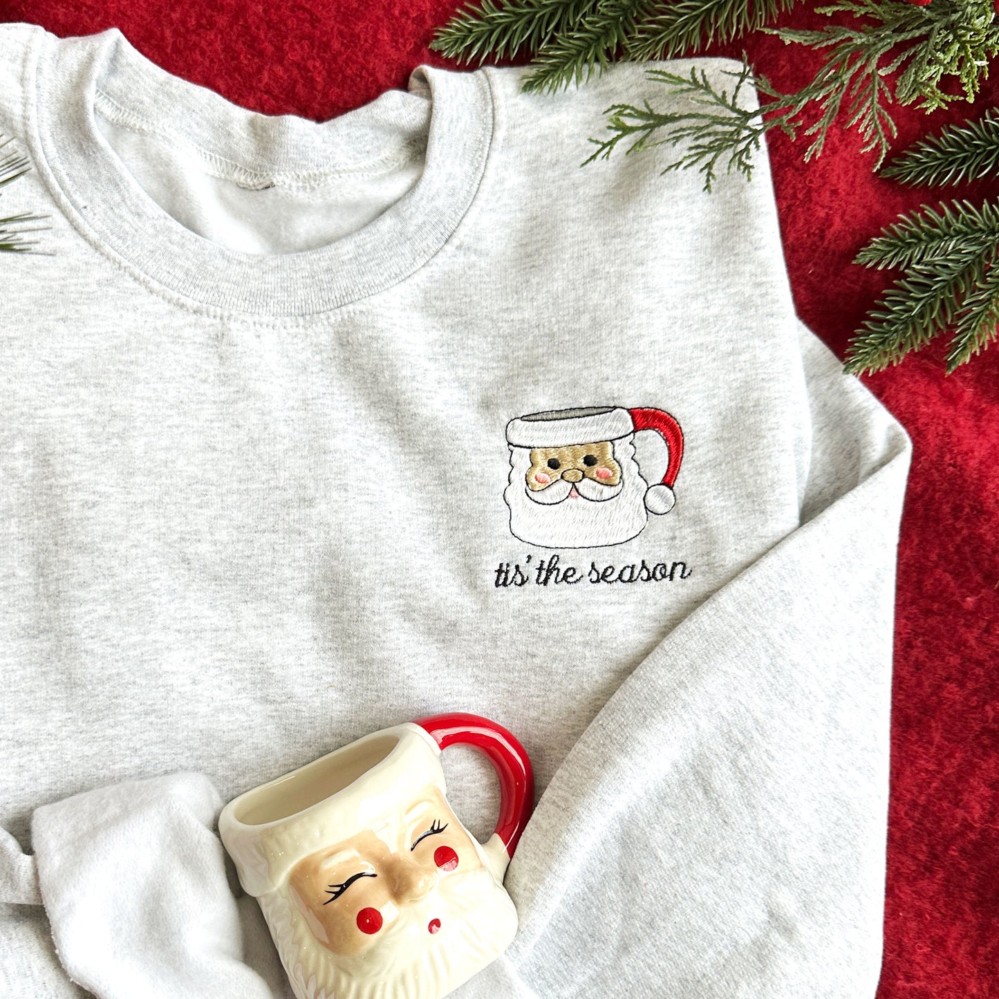 Up close image of a flay lay featuing an ash crewneck sweatshirt that showcases a santa mug embroidered design and the text tis the season underneath..