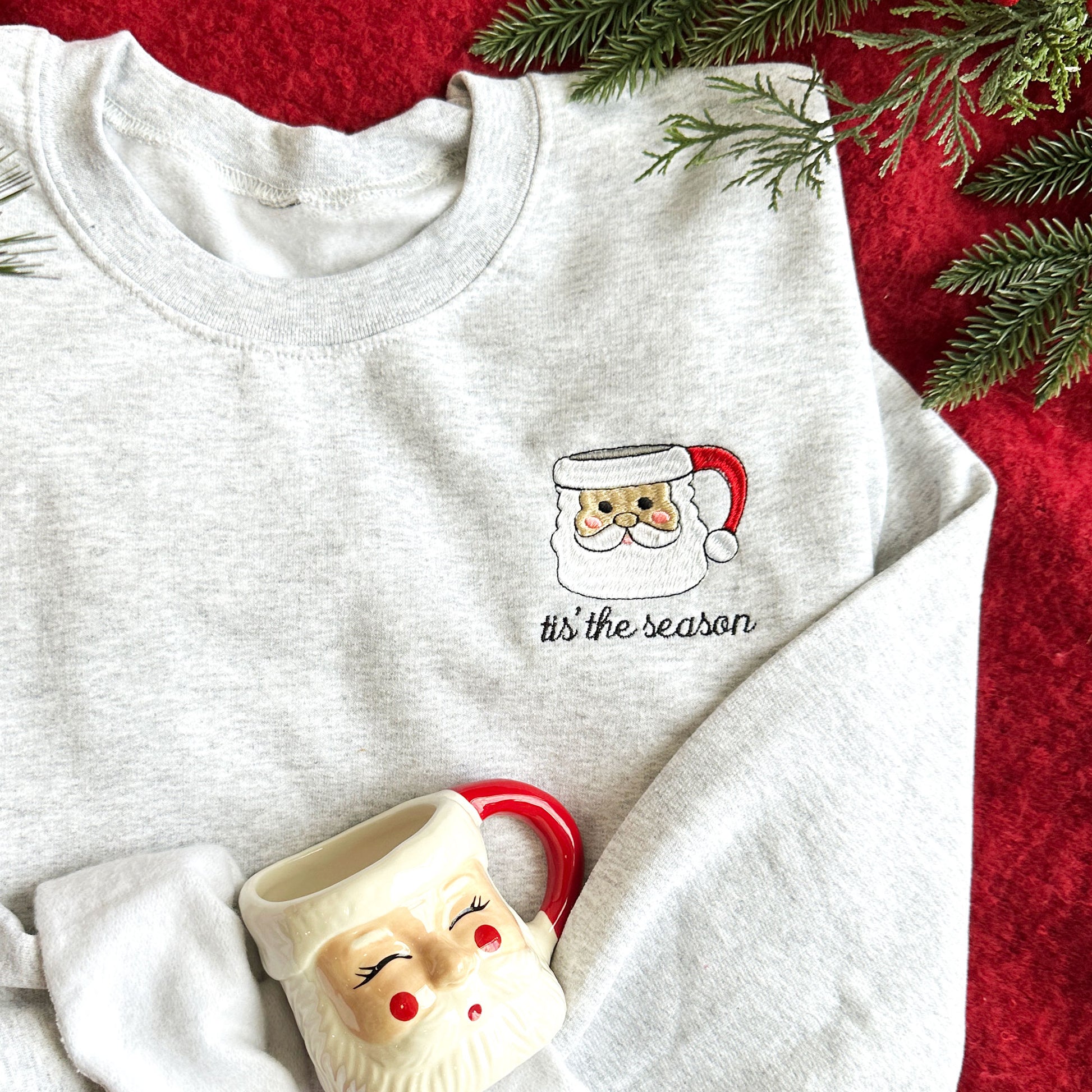 Up close image of a flay lay featuing an ash crewneck sweatshirt that showcases a santa mug embroidered design and the text tis the season underneath..