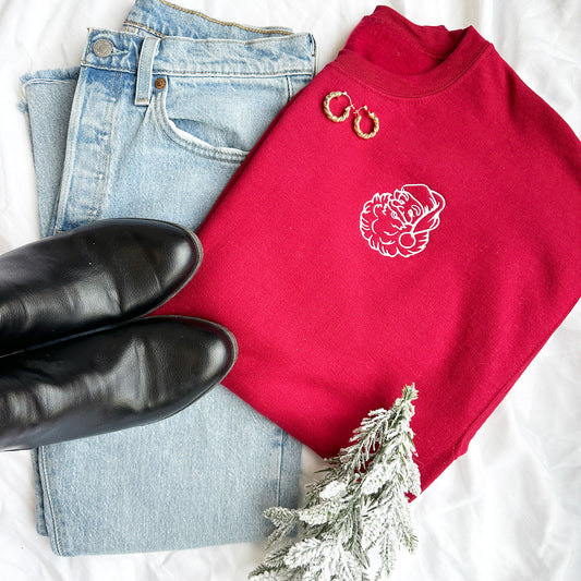 outfit flat lay featuring blue jeans, black boots, and a red crewneck sweatshirt with an outlined santa embroidered on the left chest