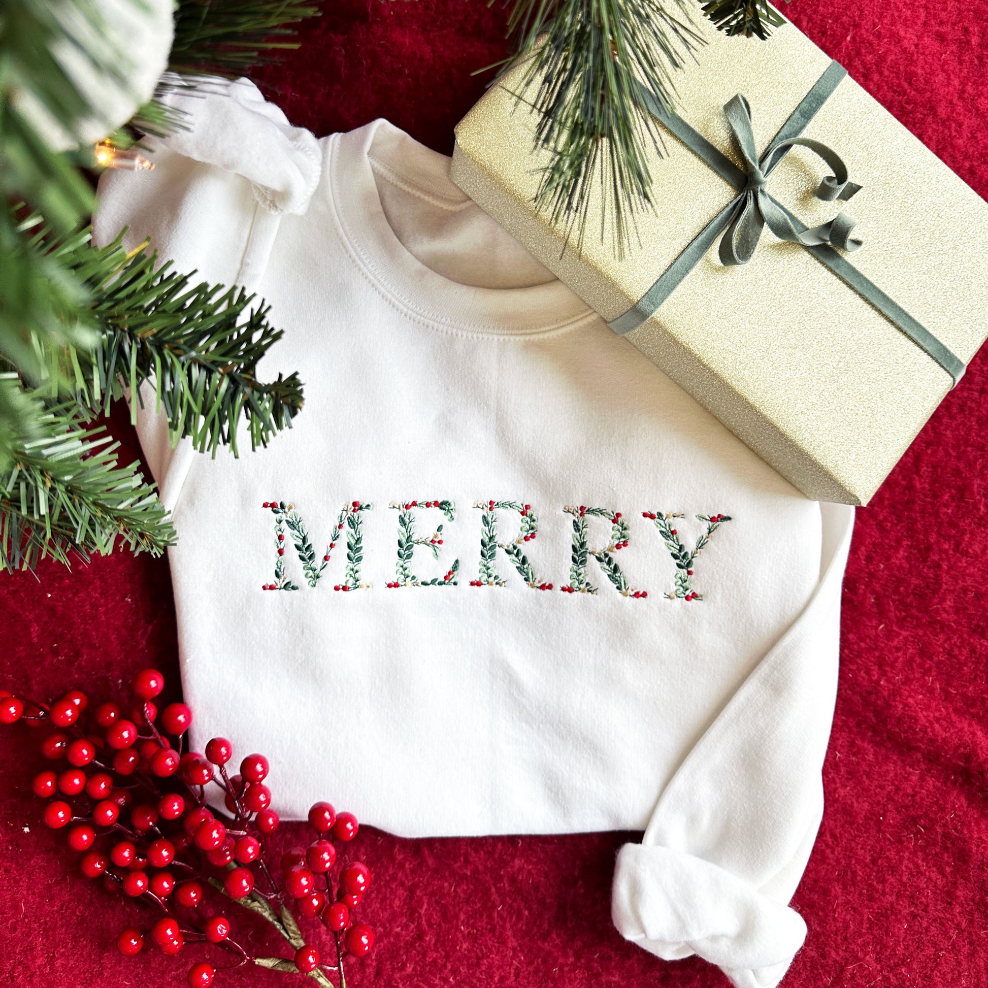 Christmas flat lay image featuring a cozy white creneck sweatshirt below a Christmas tree and next to a wrapped present. The sweatshirt has the word MERRY embroidered in floral letters and red, gold, and green threads. 