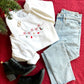 Styled flat lay image of a white crewneck sweatshirt, blue jeans, and black boots. The sweatshirt has a Christmas Garland embroidered  cross the chest with a little tree, stocking, star, ornament, and holy leaf.