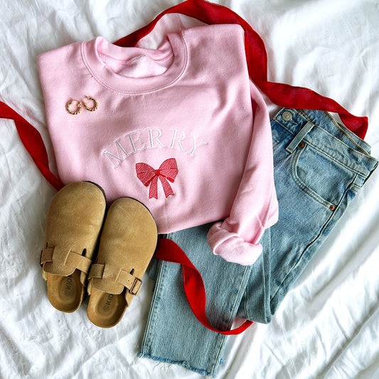 Styled flat lay photo featuring a light pink crewneck sweatshirt, blue jeans, and brown slippers. The sweatshirt features the embroidery Merry in white thread curved above a red bow.