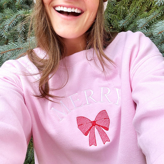 Woman taking a selfie wearing a light pink crewneck sweatshirt that has the words Merry in white embroidered above a stitched red bow.